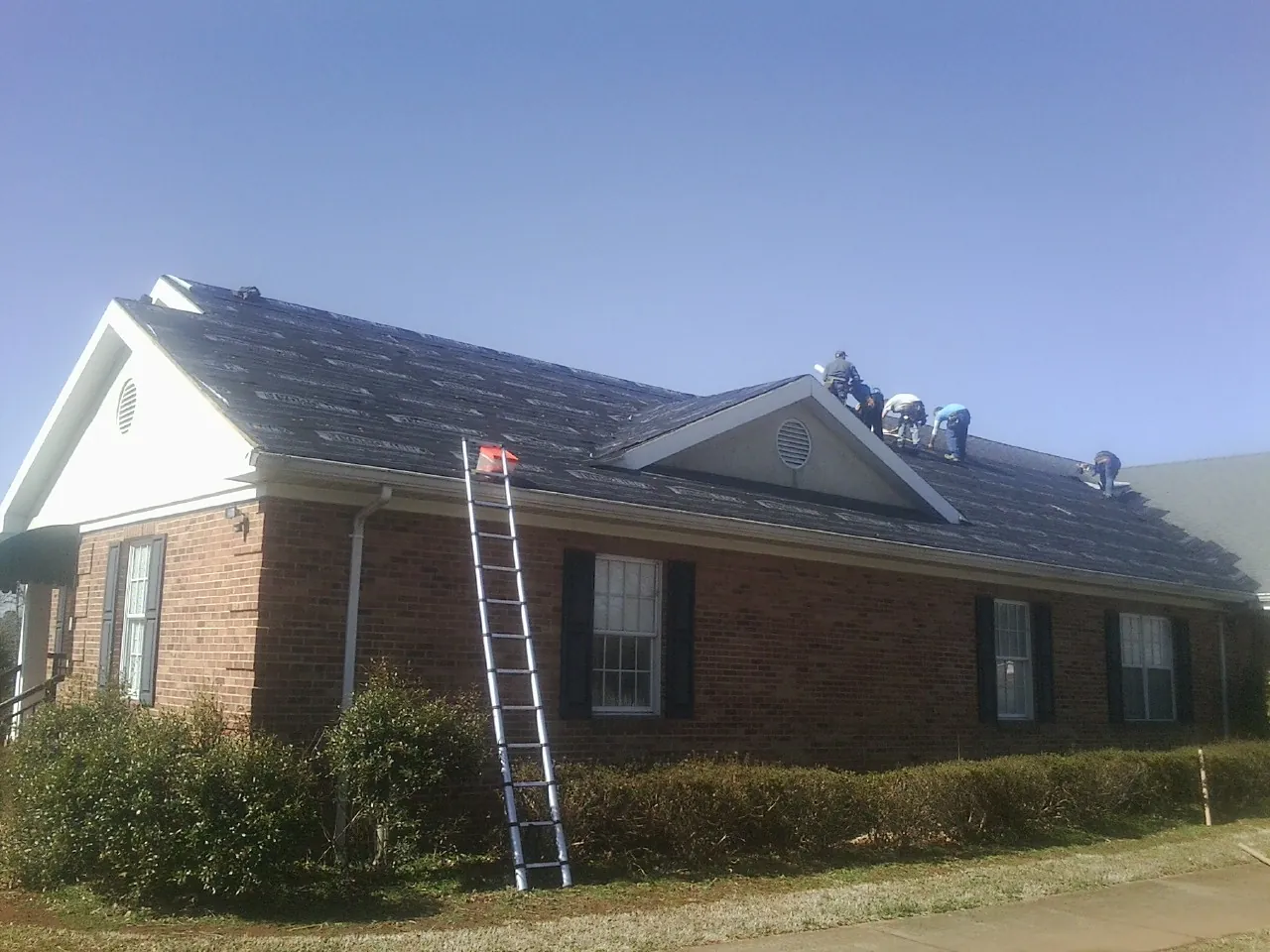 Crew tearing shingles off a brick ranch roof with a ladder set — Metal Roof Repair work by our crew in Stephenson