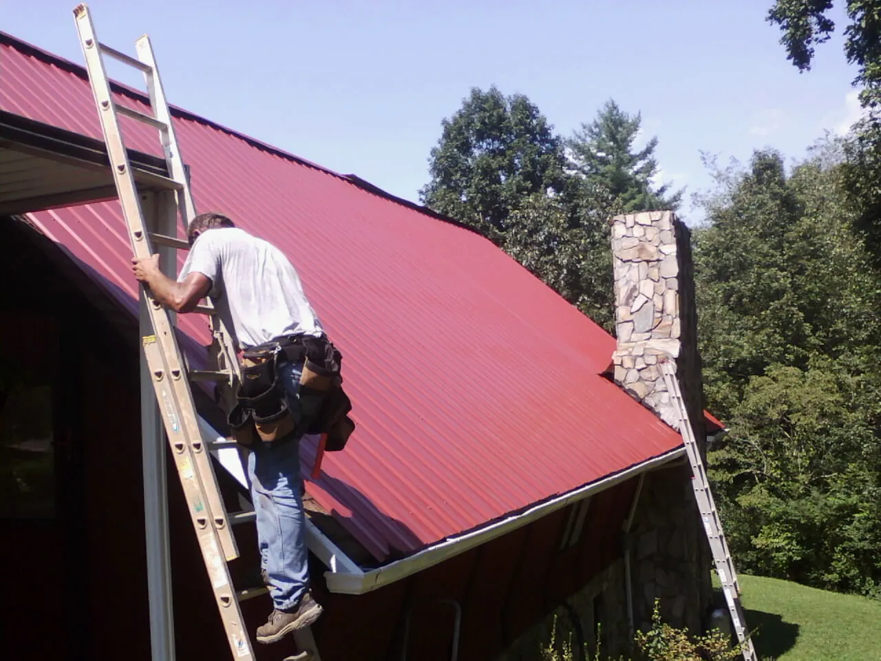 Roofer on a ladder beside a red metal roof and stone chimney — Soffit & Fascia Repair by Roofing Brothers in Stephenson